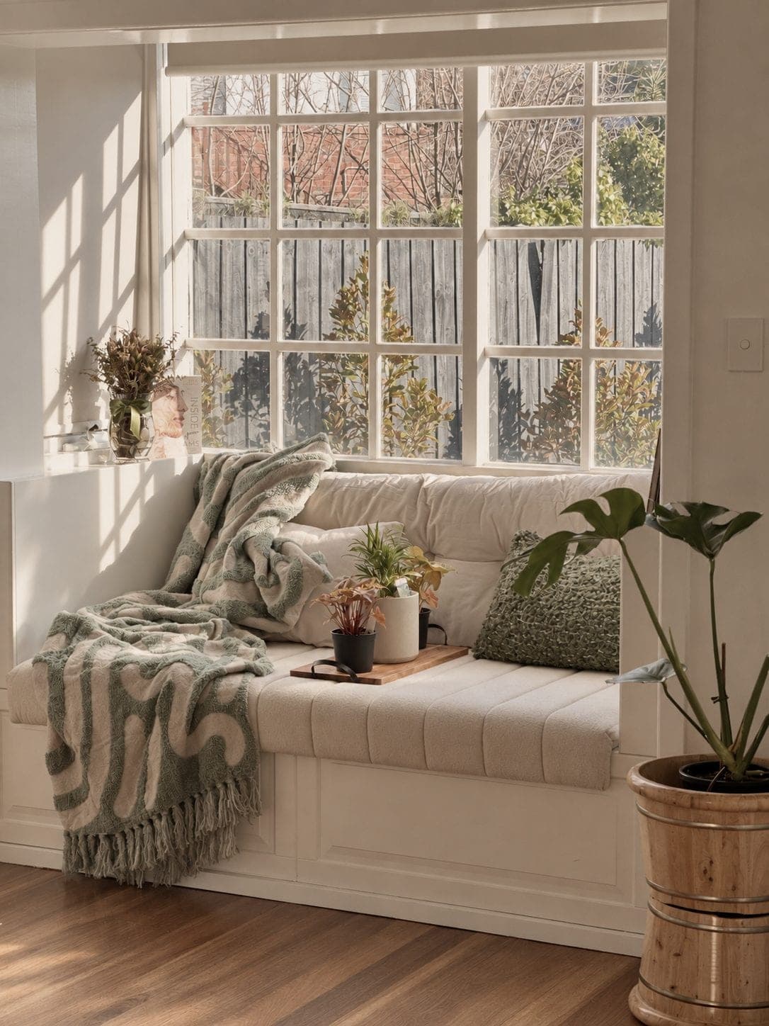 A sunlit window seat at Bötaniqa Wellbeing Studio, with a sage throw, cushions and potted plants, looking out on the garden